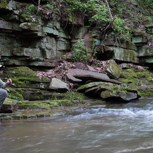 Angler kneeling on mossy shale below a rock wall to make a cast, wearing ETHIC Approach Pants in Slate