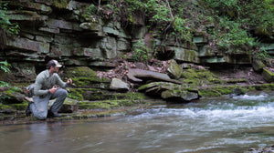 Angler kneeling on mossy shale below a rock wall to make a cast, wearing ETHIC Approach Pants in Slate
