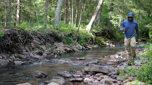 Angler walking a rocky creek bank with a fly rod, wearing the ETHIC Approach Hoodie in Blue Run with the hood up