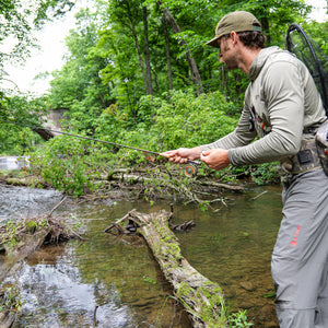Angler casting on a small wooded stream in the ETHIC Approach Hoodie in Hemlock and Approach Pants in Slate, net on his back