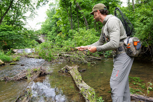Angler casting on a small wooded stream in the ETHIC Approach Hoodie in Hemlock and Approach Pants in Slate, net on his back